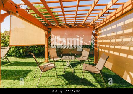 Pergolato in legno con mobili da esterno e luci sospese in un giardino che getta ombre sul terreno. Foto Stock