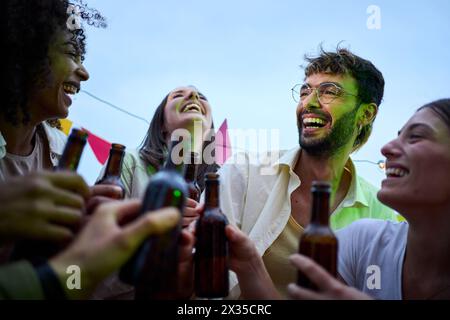 Ritratto di diversi giovani amici che ridono brindando con bottiglie di birra per festeggiare la festa Foto Stock