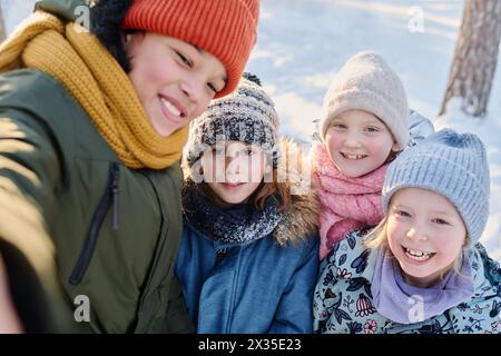 Uno dei quattro bambini felici che scattano selfie sullo smartphone mentre si trova accanto ai suoi allegri amici indossando caldi indumenti invernali Foto Stock