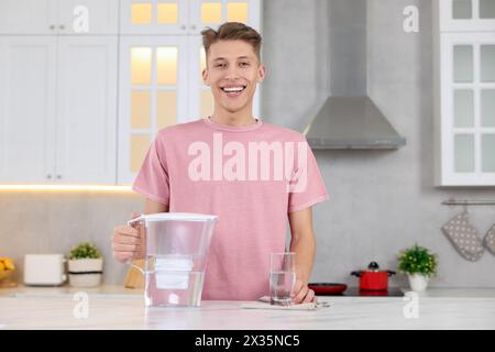 Uomo felice con caraffa filtro e bicchiere d'acqua pulita a tavola in cucina Foto Stock