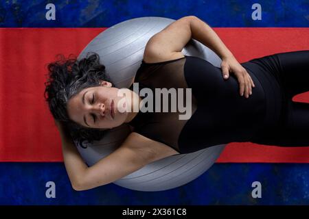 Vista dall'alto di una donna latinoamericana (38) che fa esercizi tonificanti con una palla fitness. Sfondo con tappetino rosso e pavimento blu. Studio yoga o a casa. Foto Stock