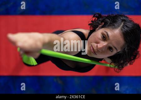 Vista dall'alto di una donna latinoamericana (38) che fa esercizi tonificanti con una fascia elastica per il fitness. Sfondo con tappetino rosso e pavimento blu. Studio yoga di concetto o. Foto Stock