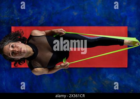 Vista dall'alto di una donna latinoamericana (38) che fa esercizi tonificanti con una fascia elastica per il fitness. Sfondo con tappetino rosso e pavimento blu. Studio yoga di concetto o. Foto Stock