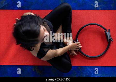 Vista dall'alto della donna latinoamericana (38) che fa esercizi tonificanti con l'anello di esercizio sul tappetino rosso per il fitness. Fondo blu. Studio yoga Foto Stock