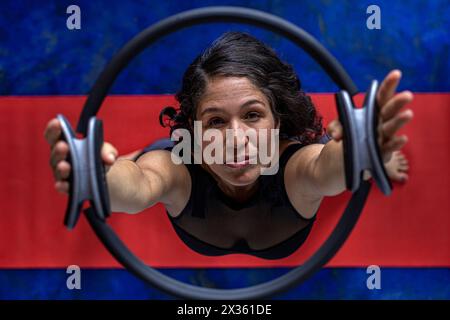 Vista dall'alto della donna latinoamericana (38) che fa esercizi tonificanti con l'anello di esercizio sul tappetino rosso per il fitness. Fondo blu. Studio yoga Foto Stock