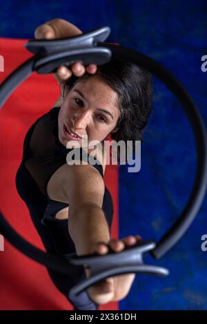 Vista dall'alto della donna latinoamericana (38) che fa esercizi tonificanti con l'anello di esercizio sul tappetino rosso per il fitness. Fondo blu. Studio yoga Foto Stock