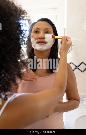 Le donne birazziali mature e le figlie usano maschere facciali, guardando allo specchio Foto Stock