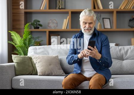 Un allegro anziano uomo caucasico seduto comodamente su un divano grigio utilizzando uno smartphone in un soggiorno ben arredato con piante verdi. Foto Stock
