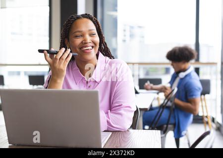 Una donna birazziale con trecce parla al telefono, lavora al computer portatile in un ufficio moderno Foto Stock