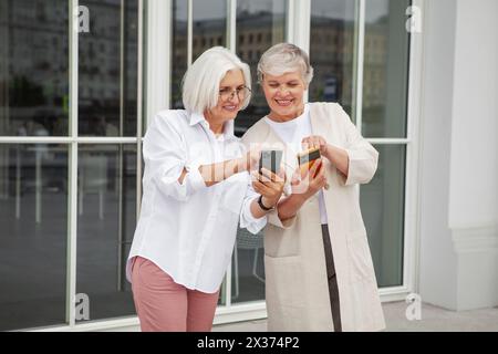 Due gioiose donne anziane in piedi fuori dall'edificio, sorridenti scambiano i dati di contatto con gli smartphone, indossano un elegante abbigliamento casual e si godono il calore Foto Stock