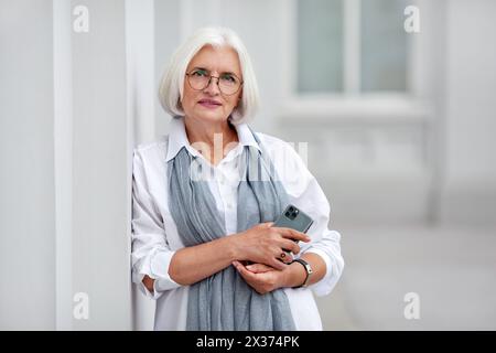 Donna d'affari matura, bella e professionale, con smartphone, felice, fiduciosa, imprenditrice modello positiva in piedi all'aperto per strada, donna senior wi Foto Stock