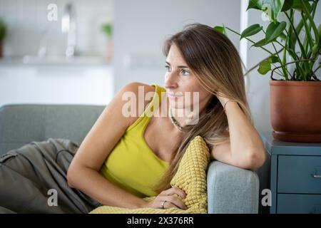 Donna rilassata seduta sul divano che trascorre il tempo pigro a casa guardando la finestra con calma emozione. Vita lenta Foto Stock