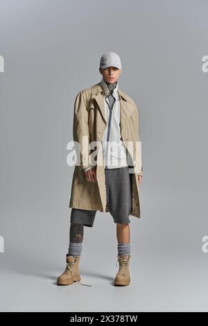 Un giovane uomo tatuato mette in posa una misteriosa veste con un trench e un cappello sullo sfondo grigio di uno studio. Foto Stock