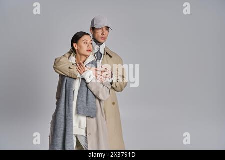 Un giovane uomo e una donna alla moda che indossano un trench si affiancano in uno studio con sfondo grigio. Foto Stock