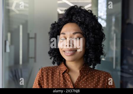 Un ritratto di una giovane donna serena che gode di un momento di pace a occhi chiusi. Sorridente dolcemente, si trova all'interno in un ambiente professionale, irradiando un'atmosfera calma e composta. Foto Stock