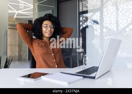 Una giovane donna d'affari nera gode di un momento di relax alla sua scrivania all'interno di un ufficio moderno. Sorride, si piega indietro con le mani dietro la testa, vicino al suo portatile. Foto Stock