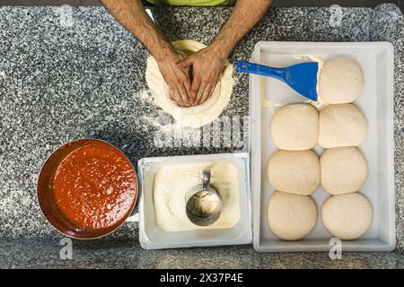 l'uomo dà la forma all'impasto della pizza, agli ingredienti e alle palline di impasto sul tavolo, vista dall'alto verso il basso. Foto di alta qualità Foto Stock