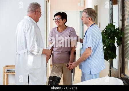 Una paziente anziana che parla con i medici nella sala clinica. Donna che indossa la schiavitù, stecca medica, ginocchio di sostegno sulla gamba, che comunica con il medico curante in un ospedale. Firmare i documenti dell'assicurazione Foto Stock