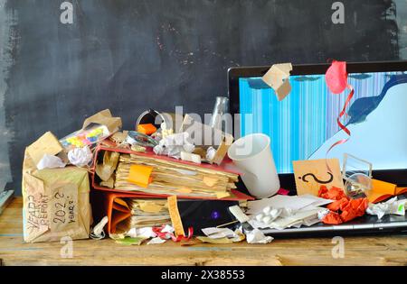 Luogo di lavoro disordinato, ufficio caotico, sovraccarico di lavoro, burocrazia, concetto di burocrazia con una scrivania da ufficio grungy, computer portatile difettoso e varie forniture per ufficio Foto Stock