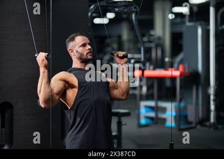 Sportivo attento che indossa una canotta nera, per allenarsi in palestra per bicipiti forti. Ritratto di muscoloso ragazzo barbuto caucasico che tira i cavi dell'allenamento A. Foto Stock