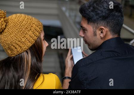 Due giovani latinoamericani in piedi con le spalle a parlare, commenta qualcosa che ha visto sul suo cellulare. Indossa un cappello giallo A. Foto Stock