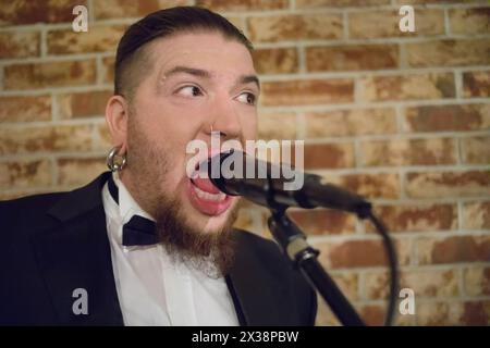 L'uomo con la barba canta nel microfono sul palco con un muro di mattoni nel club Foto Stock