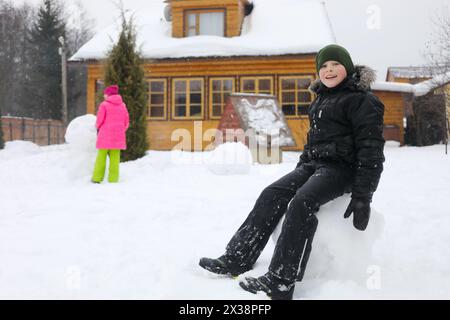 Il ragazzo siede in una grande palla di neve, la ragazza fa un pupazzo di neve vicino a una casa di campagna in legno durante le nevicate del giorno invernale, la ragazza è fuori fuoco Foto Stock