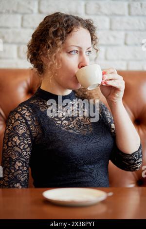 Donna in abito nero siede sul divano al tavolo bevendo un caffè nel caffè. Foto Stock