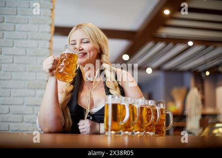 Una donna bionda sorridente sta tenendo spikelets in una mano e va a bere birra dalla tazza in un'altra al bancone del bar nel caffè. Foto Stock