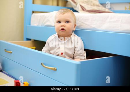 Il piccolo bel ragazzo si siede in un grande cassetto blu nella camera dei bambini a casa Foto Stock