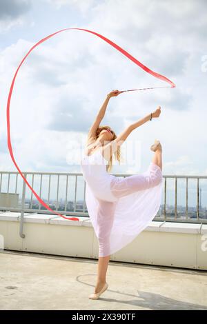 Bella donna in bianco con le mani alzate con il nastro rosso riccio in piedi su una gamba sul tetto di un edificio a più piani Foto Stock