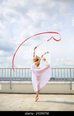 Bella ginnasta bianca con mani rialzate con nastro rosso riccio in piedi su una gamba sul tetto di un edificio a più piani Foto Stock