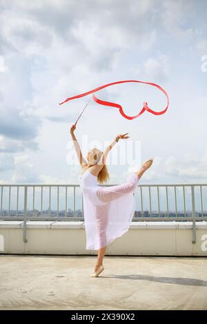 Ginnasta in bianco con mani rialzate con nastro rosso riccio in piedi su una gamba sul tetto di un edificio a più piani Foto Stock