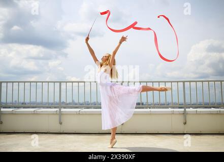 Bella ginnasta in bianco con mani alzate con nastro rosso riccio in piedi su una gamba sul tetto di un edificio a più piani Foto Stock
