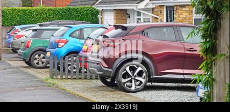Fila di colori assortiti su auto a motore berlina parcheggiate in spazi anteriori con giardino all'esterno di case private strada residenziale Essex Inghilterra Regno Unito Foto Stock
