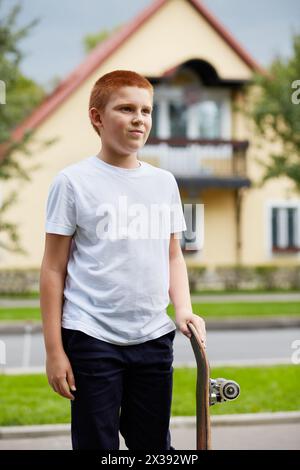 Ritratto di un ragazzo dai capelli rossi con skateboard all'aperto contro casa. Foto Stock