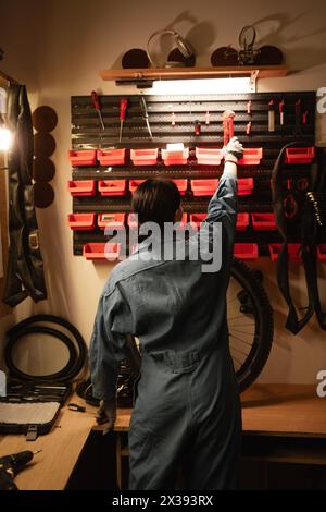Vista posteriore di un meccanico di una bicicletta bruna che lavora nel suo garage, davanti a una parete con attrezzi e attrezzature per le riparazioni Foto Stock
