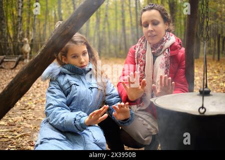 Donna e figlia siedono vicino al fuoco con la pentola appesa nella foresta asciutta autunnale Foto Stock