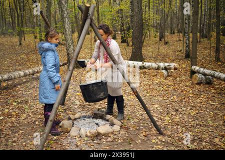 La donna rimuove il vaso appeso e la figlia la guarda vicino al falò nella foresta autunnale Foto Stock