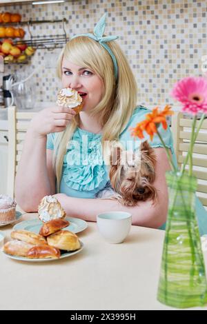 Una donna sfarzosa sorridente che tiene in mano un piccolo cane si siede a mangiare dolci fatti in casa a tavola in cucina. Foto Stock