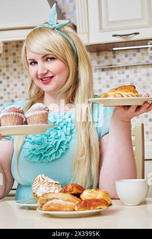 Una donna sorridente e grintosa si siede tenendo due piatti con dolci fatti in casa a tavola in cucina. Foto Stock