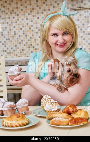 Una donna sfarzosa sorridente che tiene in mano un piccolo cane si siede a mangiare dolci fatti in casa a tavola in cucina. Foto Stock