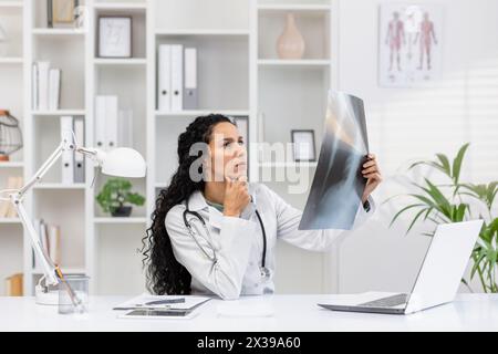 Medico ispanico mirato che esamina una pellicola radiografica mentre contempla la diagnostica in un moderno studio medico ben attrezzato. Foto Stock