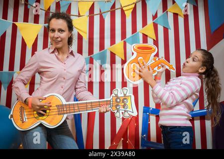 La bambina con sua madre sta suonando la chitarra e il sassofono vicino al muro a strisce. Foto Stock