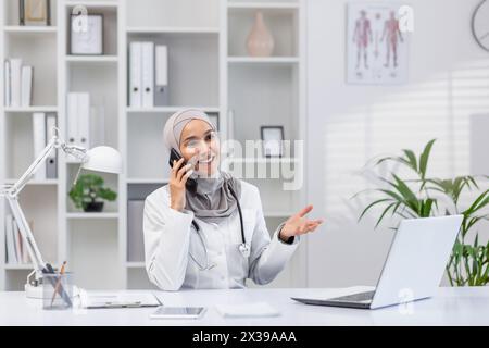 Una donna professionale e allegra che indossa un hijab si impegna in una conversazione telefonica mentre è seduta alla sua scrivania in un ufficio della clinica ben illuminato, mostrando un atteggiamento amichevole. Foto Stock