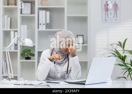 Una donna musulmana in un hijab seduta alla scrivania del suo ufficio, sentendosi stressata e mostrando segni di mal di testa mentre si lavora su un notebook. Concetto di stress sul posto di lavoro e assistenza sanitaria. Foto Stock