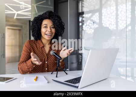 Una allegra donna afroamericana si impegna con il suo pubblico online mentre registra un podcast in un ambiente d'ufficio luminoso e moderno. Foto Stock