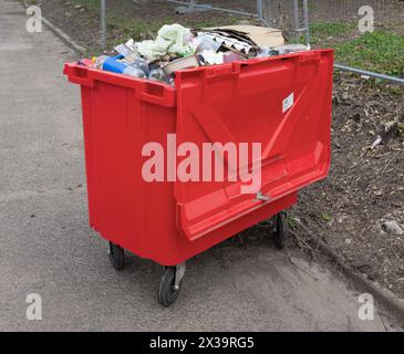 Grande bidone rosso pieno di rifiuti lungo la strada accanto a un terreno di sviluppo recintato Foto Stock