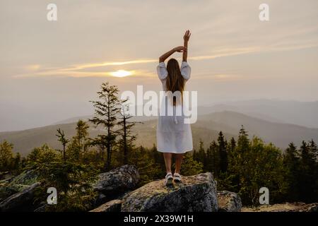 Una donna in piedi trionfalmente sulla cima di una montagna, tenendo le braccia in alto per celebrare il suo successo e la vista mozzafiato. Foto Stock