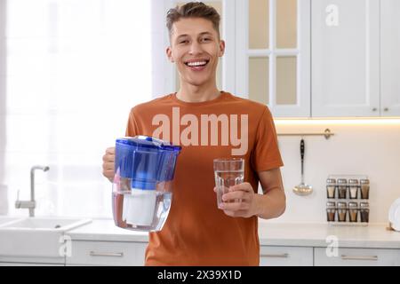 Uomo felice con caraffa filtrante e bicchiere d'acqua pulita in cucina Foto Stock